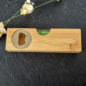May include: A wooden bottle opener with a built-in level. The opener is made of light brown bamboo and has a silver metal bottle opener.