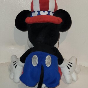 Op de afbeelding: Een pluche Mickey Mouse speelgoed dat een rood, wit en blauwe patriottische hoed draagt. Het speelgoed zit op een wit oppervlak.