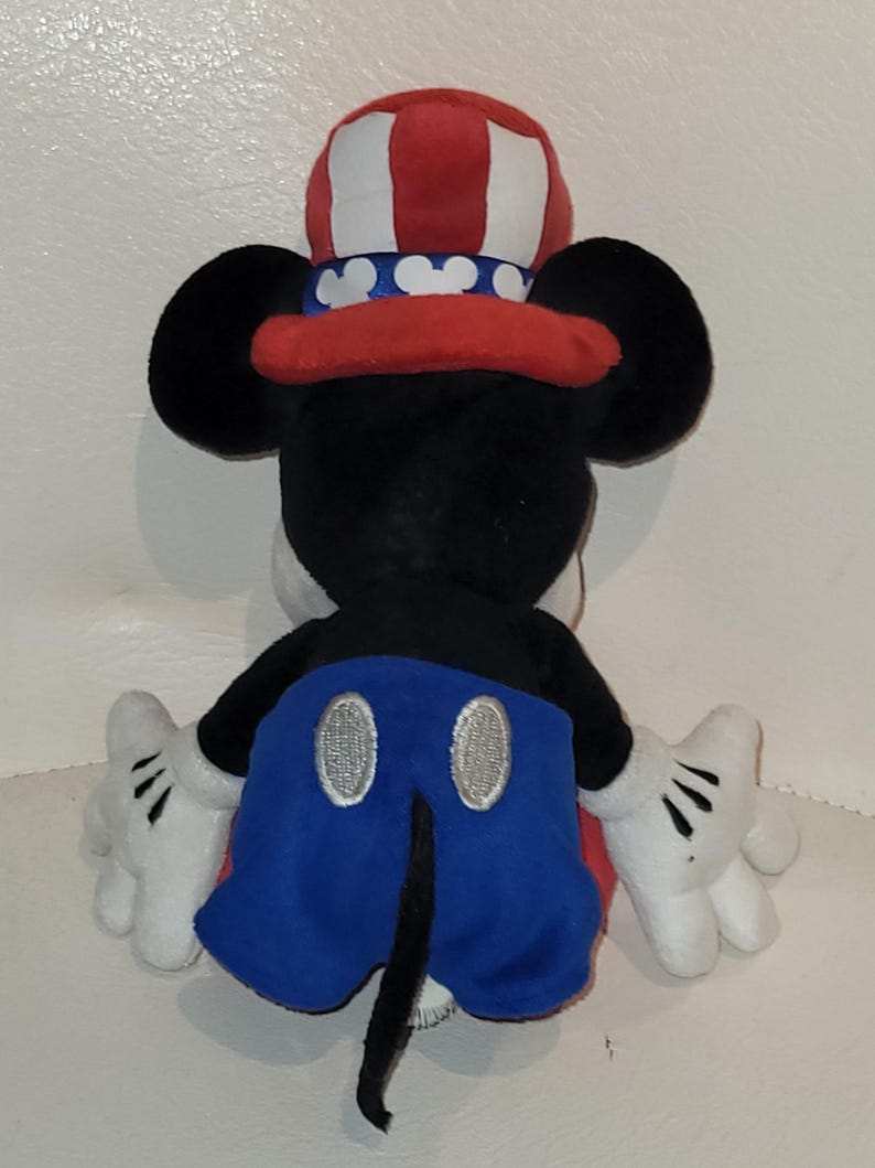 Op de afbeelding: Een pluche Mickey Mouse speelgoed dat een rood, wit en blauwe patriottische hoed draagt. Het speelgoed zit op een wit oppervlak.