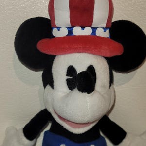 Op de afbeelding: Een pluche Mickey Mouse speelgoed dat een rood, wit en blauwe patriottische hoed draagt. De hoed heeft witte strepen en een blauwe band met witte Mickey Mouse oren. Mickey draagt een blauwe broek met witte knopen.