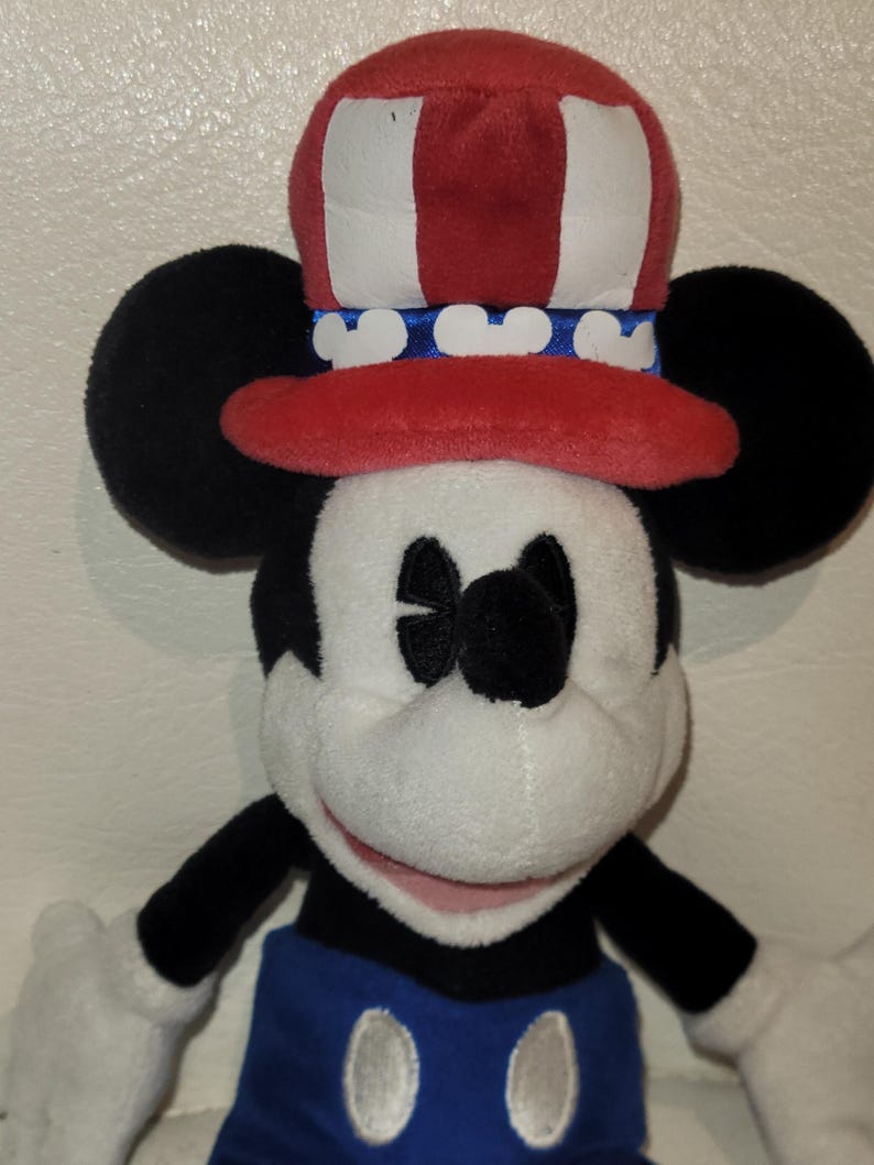 Op de afbeelding: Een pluche Mickey Mouse speelgoed dat een rood, wit en blauwe patriottische hoed draagt. De hoed heeft witte strepen en een blauwe band met witte Mickey Mouse oren. Mickey draagt een blauwe broek met witte knopen.