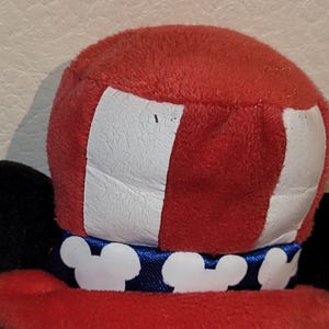 Op de afbeelding: Een zwarte en witte pluche Mickey Mouse die een rood, wit en blauwe hoed draagt met een blauw lint en witte Mickey Mouse-oren. De hoed heeft een wit en rood gestreept patroon.