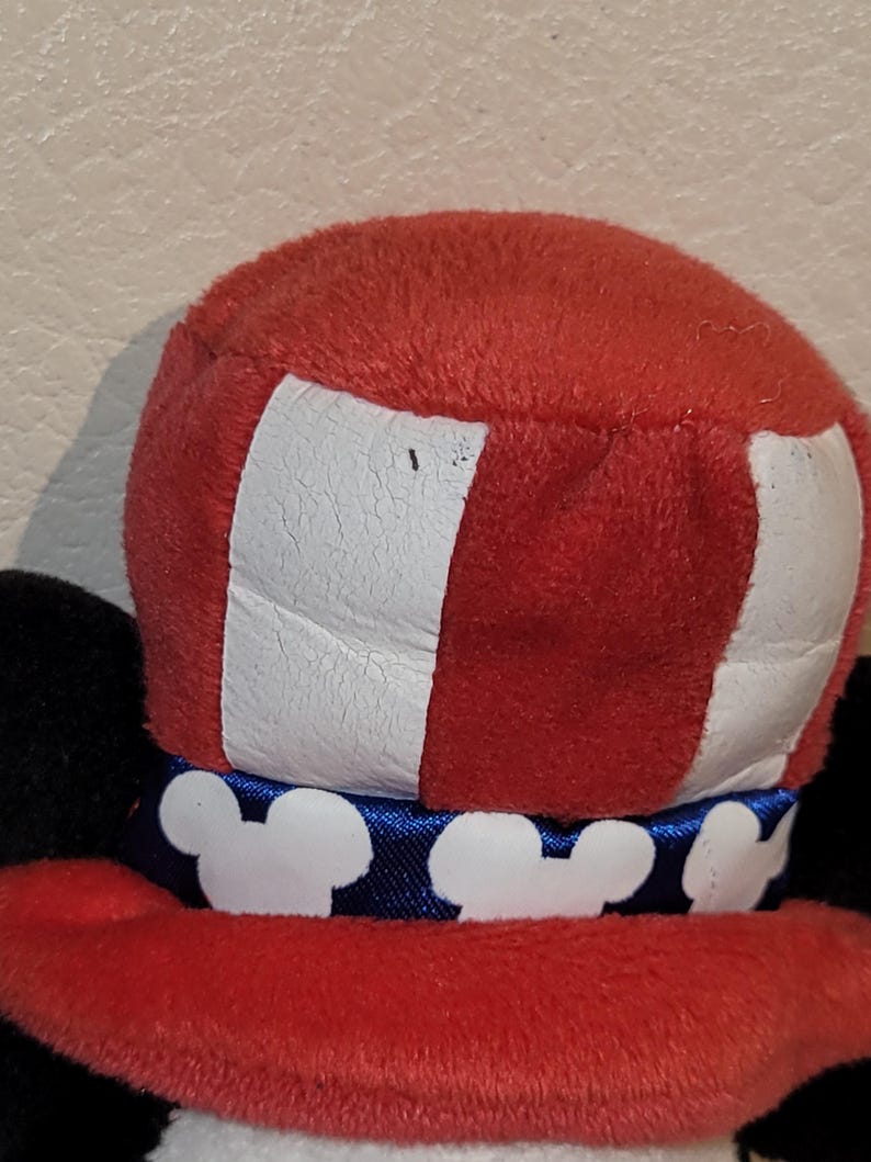 Op de afbeelding: Een zwarte en witte pluche Mickey Mouse die een rood, wit en blauwe hoed draagt met een blauw lint en witte Mickey Mouse-oren. De hoed heeft een wit en rood gestreept patroon.