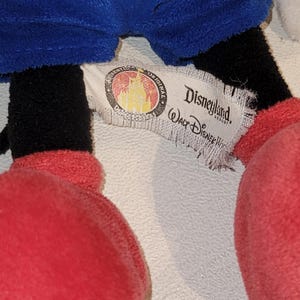 Op de afbeelding: Een close-up van het label van een Mickey Mouse pluche speelgoed. Het label is wit met zwarte tekst die "Disneyland Resort" en "Walt Disney World" zegt. Het label heeft ook een rood en gouden logo met een kasteel en sterren.