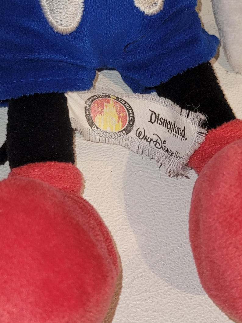 Op de afbeelding: Een close-up van het label van een Mickey Mouse pluche speelgoed. Het label is wit met zwarte tekst die "Disneyland Resort" en "Walt Disney World" zegt. Het label heeft ook een rood en gouden logo met een kasteel en sterren.