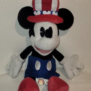 Op de afbeelding: Een pluche Mickey Mouse die een rood, wit en blauwe hoed draagt met een blauw-wit gestreepte band. Mickey draagt een blauwe broek met een zilveren knoop en rode schoenen. De pluche zit op een wit oppervlak.