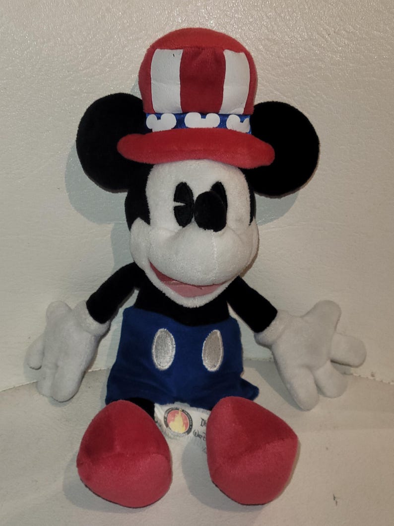 Op de afbeelding: Een pluche Mickey Mouse die een rood, wit en blauwe hoed draagt met een blauw-wit gestreepte band. Mickey draagt een blauwe broek met een zilveren knoop en rode schoenen. De pluche zit op een wit oppervlak.
