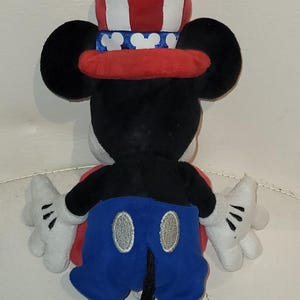 Op de afbeelding: Een pluche Mickey Mouse speelgoed dat een rood, wit en blauwe hoed draagt met sterren en strepen. Mickey draagt een blauwe broek met witte knopen en zwarte schoenen.