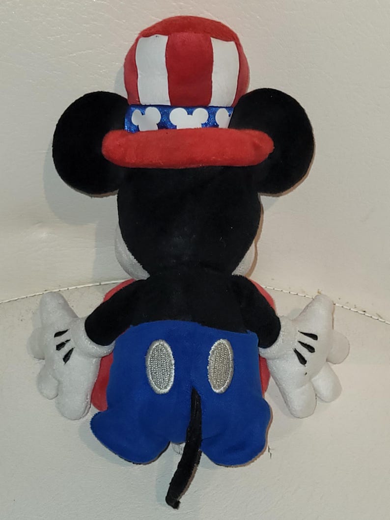 Op de afbeelding: Een pluche Mickey Mouse speelgoed dat een rood, wit en blauwe hoed draagt met sterren en strepen. Mickey draagt een blauwe broek met witte knopen en zwarte schoenen.