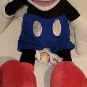 Op de afbeelding: Een pluche Mickey Mouse speelgoed met zwarte armen en benen, blauwe shorts met witte knopen en rode schoenen. Het speelgoed heeft een wit gezicht met een roze mond en zwarte ogen. Het speelgoed heeft een Disney Store-label aan zijn been.