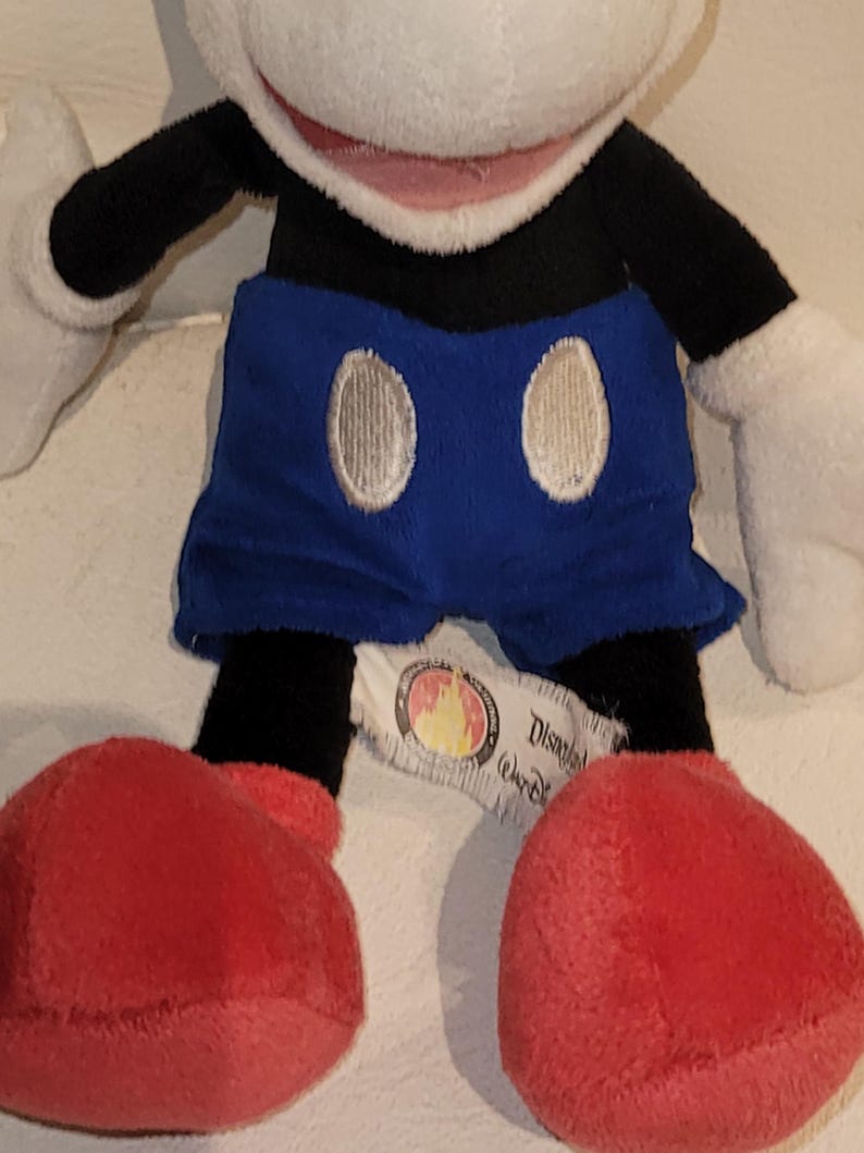 Op de afbeelding: Een pluche Mickey Mouse speelgoed met zwarte armen en benen, blauwe shorts met witte knopen en rode schoenen. Het speelgoed heeft een wit gezicht met een roze mond en zwarte ogen. Het speelgoed heeft een Disney Store-label aan zijn been.