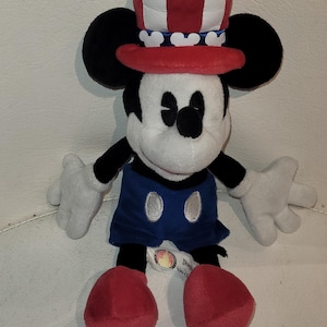 Op de afbeelding: Een pluche Mickey Mouse met een rood, wit en blauwe hoed met een patriottisch thema. Het pluche speelgoed zit met gekruiste benen en heeft een blauwe en witte outfit.