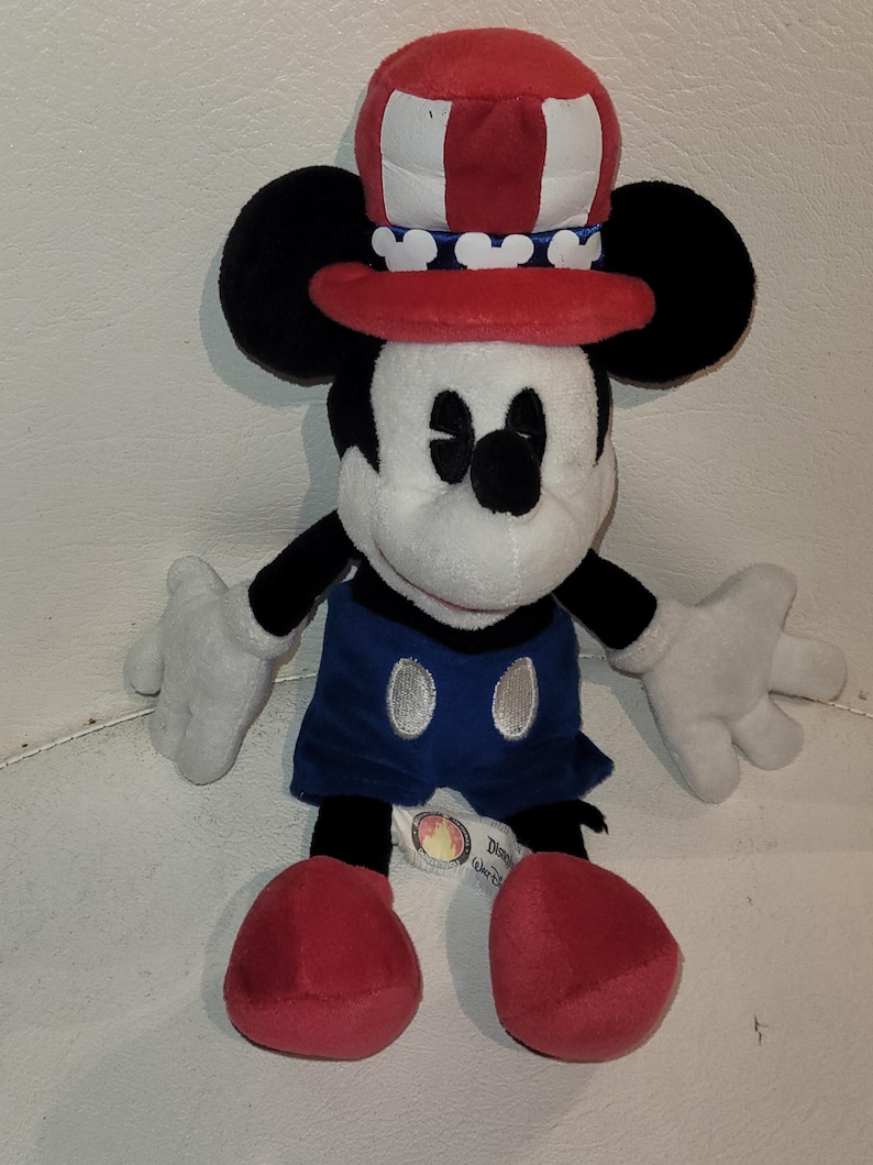 Op de afbeelding: Een pluche Mickey Mouse met een rood, wit en blauwe hoed met een patriottisch thema. Het pluche speelgoed zit met gekruiste benen en heeft een blauwe en witte outfit.