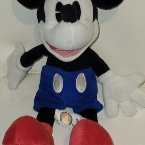 Op de afbeelding: Een pluche Mickey Mouse speelgoed dat een rood, wit en blauwe hoed draagt en blauwe shorts met zilveren knopen. Het speelgoed heeft een grote glimlach en zit met gekruiste benen.