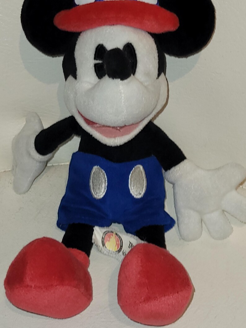 Op de afbeelding: Een pluche Mickey Mouse speelgoed dat een rood, wit en blauwe hoed draagt en blauwe shorts met zilveren knopen. Het speelgoed heeft een grote glimlach en zit met gekruiste benen.