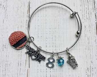 May include: A silver-toned adjustable bangle bracelet with basketball-themed charms. Charms include a basketball, the words "BASKETBALL MOM", the number 8, a blue bead, and a basketball net. The basketball charm is brown and black.