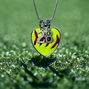 Puede incluir: Un collar con un dije de softball amarillo y un dije de plata con el número 28. El collar está sobre un fondo de hierba verde.
