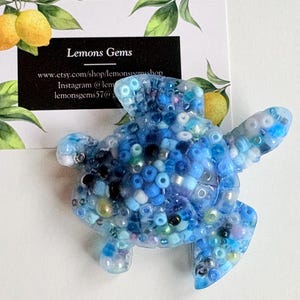 May include: A blue resin turtle figurine filled with various shades of blue beads. The turtle has a textured surface and is displayed on a white background with a business card that reads "Lemons Gems".