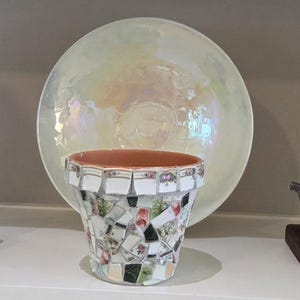 May include: A terracotta flower pot decorated with a mosaic of broken ceramic pieces featuring floral patterns and gold trim. Behind the pot sits a large, round, iridescent bowl with a pearlescent finish that reflects light in soft pastel tones.