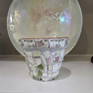 May include: A terracotta flower pot decorated with a mosaic of broken ceramic pieces. The shards feature white, green, and floral patterns with pink and blue blossoms. The pieces are set in gray grout, creating a textured surface around the exterior.