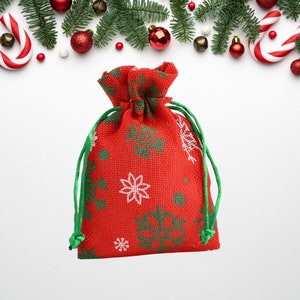 Puede incluir: Una bolsa de yute roja con un patrón de copo de nieve verde y blanco. La bolsa es perfecta para guardar pequeños regalos o favores.