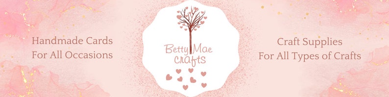 May include: A banner with a pink watercolor background and gold accents. The text reads "Handmade Cards For All Occasions" and "Craft Supplies For All Types of Crafts". The center features a logo with the text "Betty Mae crafts" and a tree design.