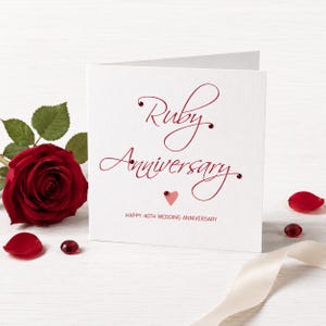 May include: A white greeting card with the text Ruby Anniversary and Happy 40th Wedding Anniversary in red script and block lettering. The card is decorated with small red gems and a pink heart, accompanied by a red rose and scattered red petals.