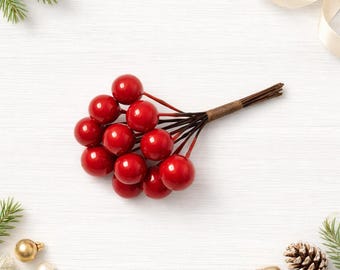 Puede incluir: Un pequeño racimo decorativo de bayas rojas brillantes unidas a finos tallos de alambre marrón. Las bayas son redondas y lisas, reunidas en la base con una atadura de papel marrón, creando un manojo compacto y uniforme de fruta artificial.