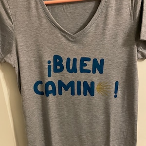May include: Gray V-neck t-shirt with the text "¡BUEN CAMINO!" in blue with a yellow scallop shell graphic.
