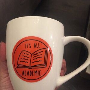 Op de afbeelding: Witte keramische mok met een fel oranje cirkelontwerp. De cirkel heeft een zwarte omtrek van een open boek en de tekst "It's All Academic".