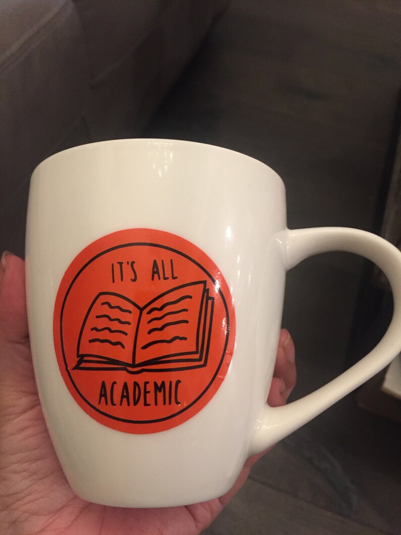 Op de afbeelding: Witte keramische mok met een fel oranje cirkelontwerp. De cirkel heeft een zwarte omtrek van een open boek en de tekst "It's All Academic".