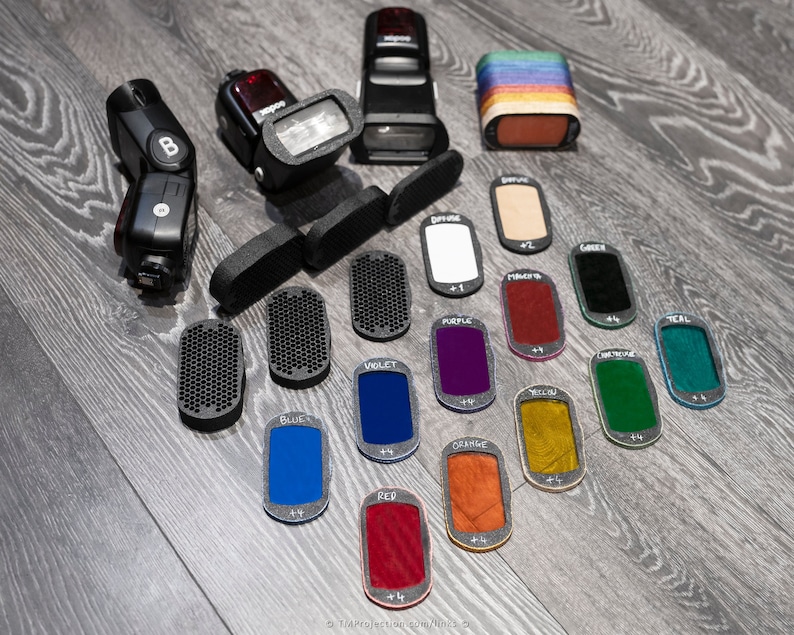 May include: A set of colored gel filters for flash photography. The filters are labeled with their colors, including red, orange, yellow, green, teal, blue, violet, and purple. The filters are arranged in a row on a wooden surface.