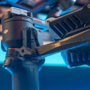 May include: Close-up of a black DJI camera gimbal with a silver control knob and a red accent. The gimbal is attached to a black and silver arm with a textured surface.
