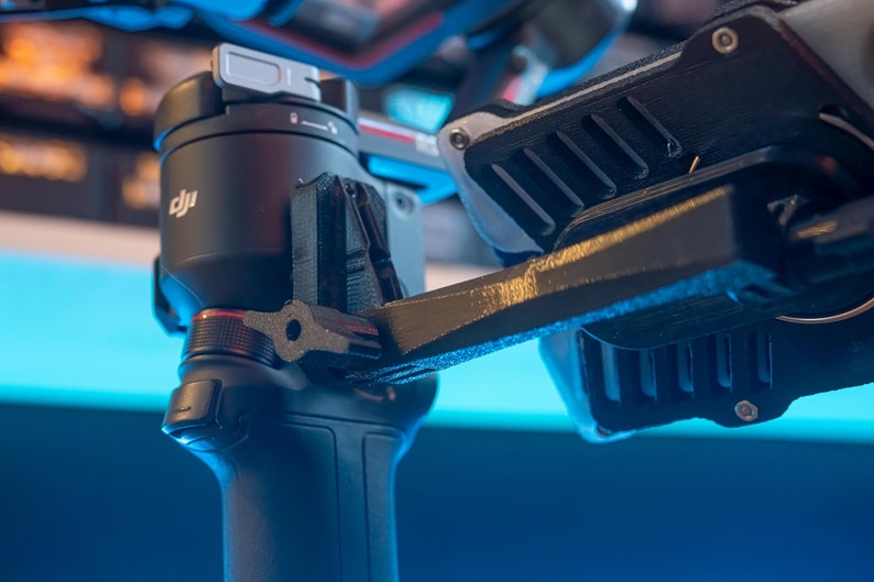 May include: Close-up of a black DJI camera gimbal with a silver control knob and a red accent. The gimbal is attached to a black and silver arm with a textured surface.