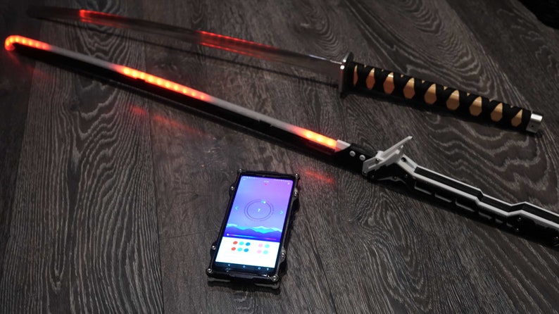 Pode incluir: Duas espadas, uma com cabo preto e dourado e a outra com cabo preto e prateado, est&atilde;o sobre uma superf&iacute;cie de madeira escura. Um smartphone com uma capa preta est&aacute; no centro da imagem, exibindo um aplicativo de sele&ccedil;&atilde;o de cores.