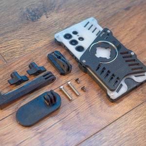 May include: A white and black phone case with a ring holder and a camera lens cover. The case comes with a variety of black plastic accessories, including a phone stand, a grip, and screws.