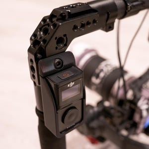 Puede incluir: Plataforma de c&aacute;mara negra con una c&aacute;mara montada y una peque&ntilde;a pantalla. La plataforma tiene m&uacute;ltiples puntos de montaje y un asa. La c&aacute;mara tiene un bot&oacute;n de encendido y el logotipo de DJI. El fondo est&aacute; borroso.