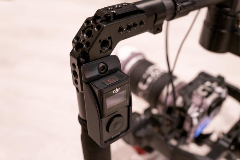 Puede incluir: Plataforma de c&aacute;mara negra con una c&aacute;mara montada y una peque&ntilde;a pantalla. La plataforma tiene m&uacute;ltiples puntos de montaje y un asa. La c&aacute;mara tiene un bot&oacute;n de encendido y el logotipo de DJI. El fondo est&aacute; borroso.