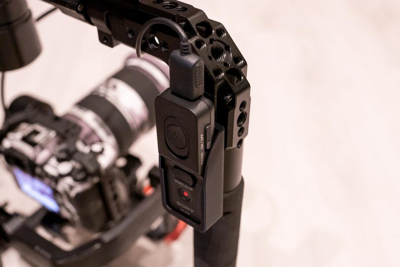 Puede incluir: Plataforma de c&aacute;mara negra con un m&oacute;dulo de control negro adjunto. El m&oacute;dulo tiene una luz roja y la palabra "SONY" impresa. La plataforma est&aacute; montada sobre una superficie blanca. El objetivo de la c&aacute;mara es visible en el fondo.