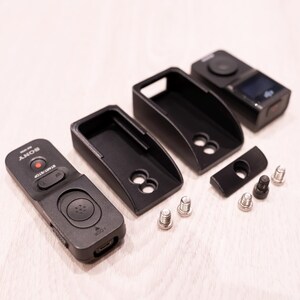 Puede incluir: Accesorios de c&aacute;mara negros, que incluyen un control remoto, fundas protectoras y hardware de montaje. El control remoto tiene un bot&oacute;n de grabaci&oacute;n rojo y la marca "SONY". Las fundas tienen un dise&ntilde;o curvo con recortes circulares.