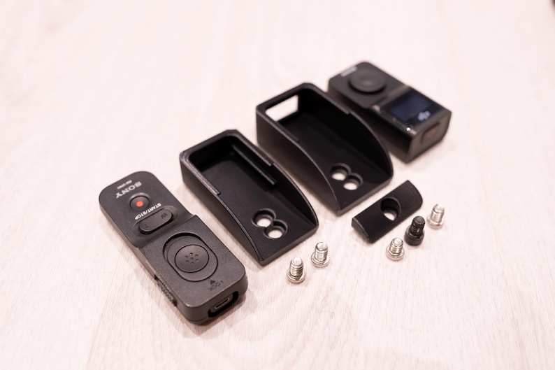 Puede incluir: Accesorios de c&aacute;mara negros, que incluyen un control remoto, fundas protectoras y hardware de montaje. El control remoto tiene un bot&oacute;n de grabaci&oacute;n rojo y la marca "SONY". Las fundas tienen un dise&ntilde;o curvo con recortes circulares.