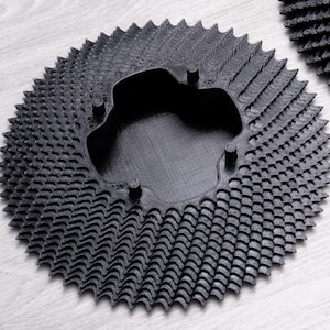 Könnte beinhalten: Ein schwarzes, kreisförmiges Zahnrad mit einem komplexen, strukturierten Design. Das Zahnrad hat eine zentrale Öffnung und zahlreiche kleine, spitze Zähne am äußeren Rand. Das Zahnrad besteht aus einem dunklen Material und liegt auf einer hellen Oberfläche.