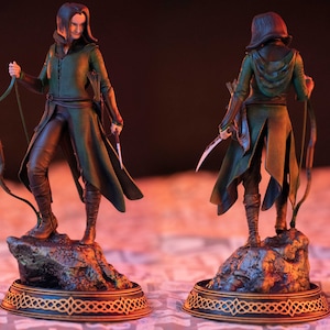 May include: A green and brown figurine of a female character with long dark hair, wearing a hooded cloak and holding a bow and arrow. The figurine is standing on a rocky base with a gold and black circular base.