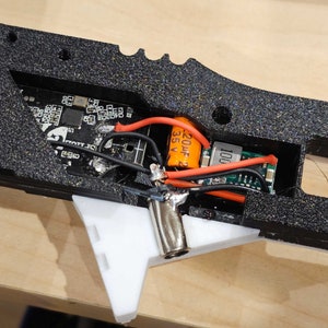 Pode incluir: Close-up do interior de um dispositivo preto impresso em 3D com um capacitor laranja etiquetado como "220uF 35V" e uma placa de circuito impresso preta com o texto "1001".
