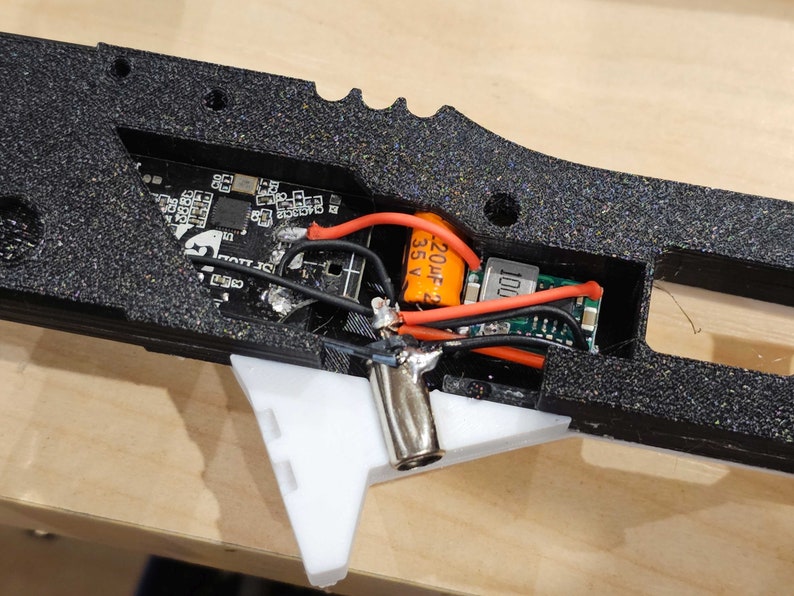 Pode incluir: Close-up do interior de um dispositivo preto impresso em 3D com um capacitor laranja etiquetado como "220uF 35V" e uma placa de circuito impresso preta com o texto "1001".