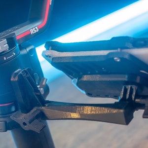 May include: A black DJI RS3 camera stabiliser with a black and grey phone mount attached to it. The stabiliser has a red and silver accent on the side. The phone mount is attached to the stabiliser with a black arm.