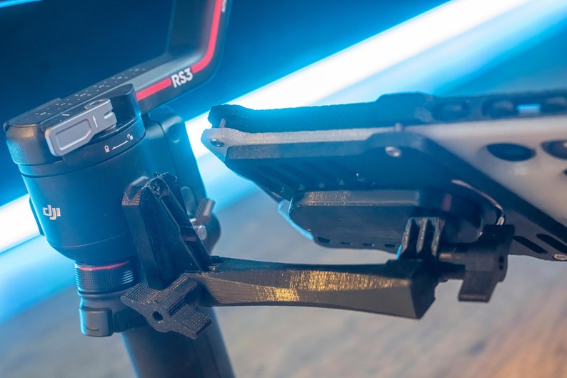 May include: A black DJI RS3 camera stabiliser with a black and grey phone mount attached to it. The stabiliser has a red and silver accent on the side. The phone mount is attached to the stabiliser with a black arm.