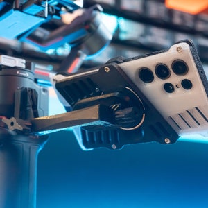 May include: A black and grey phone stabiliser with a white phone case attached. The phone case has a black camera bump with multiple lenses.