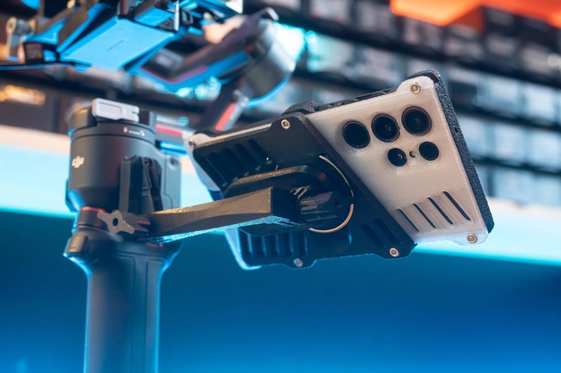 May include: A black and grey phone stabiliser with a white phone case attached. The phone case has a black camera bump with multiple lenses.