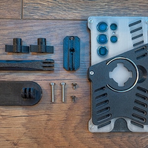 May include: A black and silver phone case with a built-in camera lens and a ring holder. The case comes with a variety of accessories, including a phone mount, screws, and a small black piece with a GoPro mount.